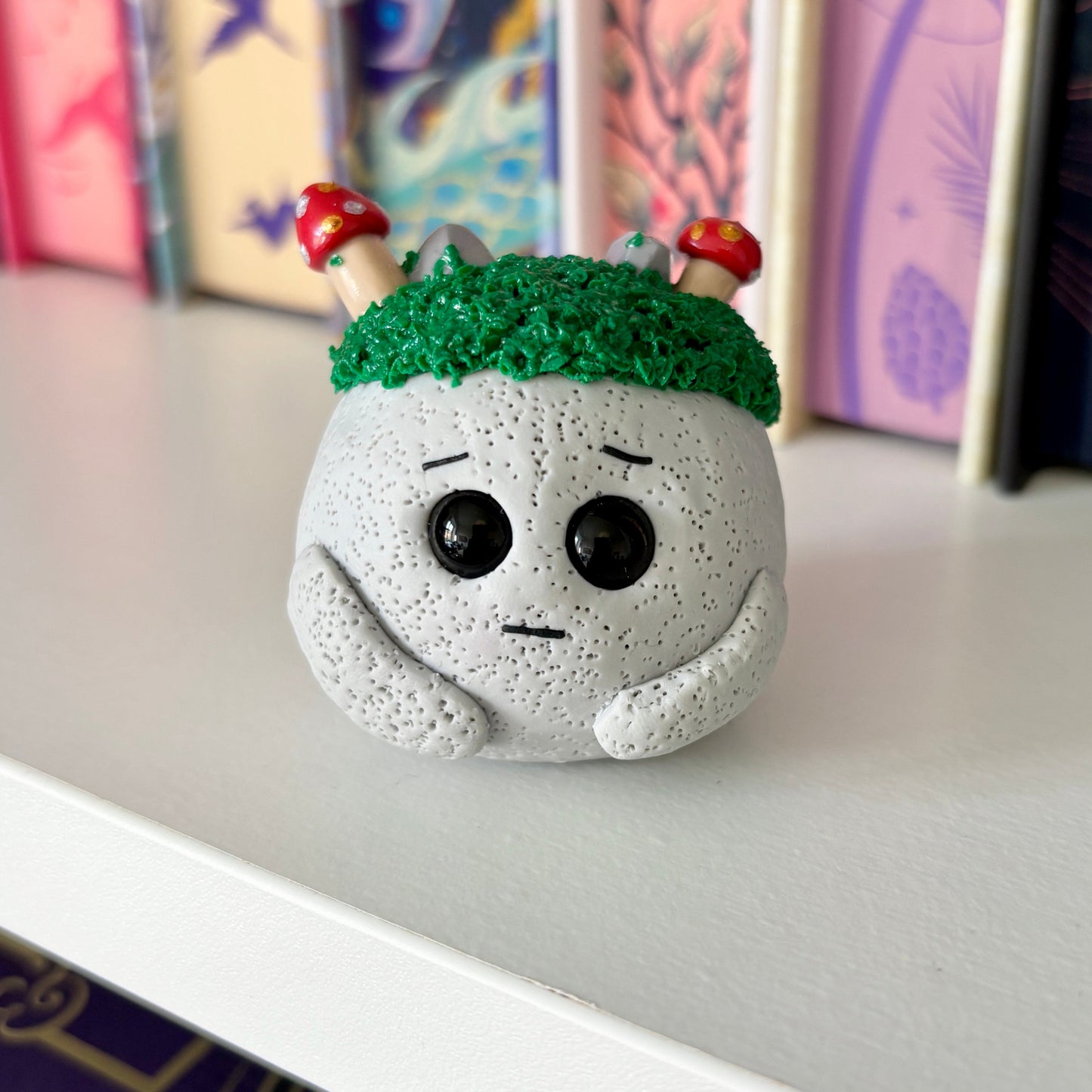 Small figurine with green moss on its head, with small red mushrooms and grey boulders on the moss. on a shelf with colorful books in the background